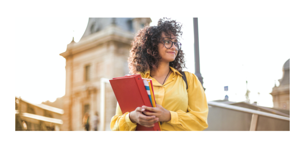 girl in yellow shirt holding red notebook in front of college building